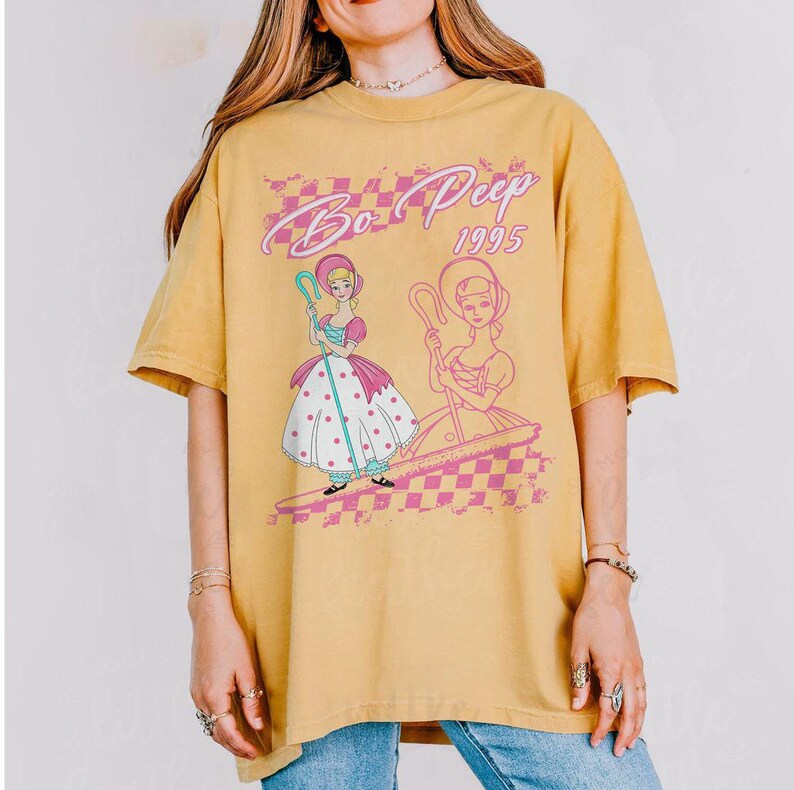 K&ouml;nnte beinhalten: Senfgelbes T-Shirt mit einer Grafik von Bo Peep aus Toy Story. Das Design umfasst zwei Illustrationen von Bo Peep, den Text "Bo Peep 1995" und ein rosa-wei&szlig;es Karomuster. Das Shirt hat kurze &Auml;rmel und eine lockere Passform.