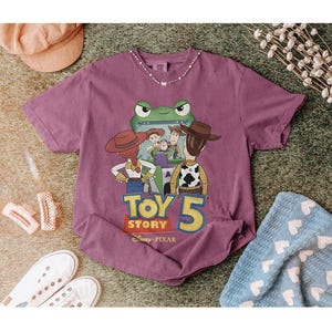 May include: A purple short-sleeved t-shirt featuring a graphic from Toy Story 5. The design includes characters like Woody and Buzz Lightyear, with the text "Toy Story 5" in yellow and white. The shirt is surrounded by accessories.