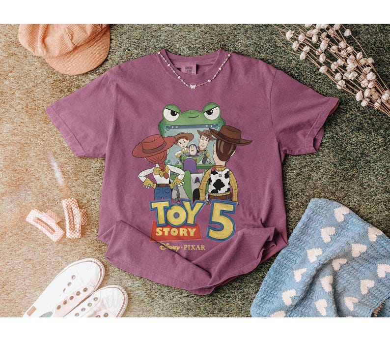 May include: A purple short-sleeved t-shirt featuring a graphic from Toy Story 5. The design includes characters like Woody and Buzz Lightyear, with the text "Toy Story 5" in yellow and white. The shirt is surrounded by accessories.