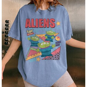 May include: A light blue t-shirt featuring the word "ALIENS" in red block letters, with the "Toy Story" logo and three green alien characters. The shirt includes the text "SINCE 1995" and star graphics.