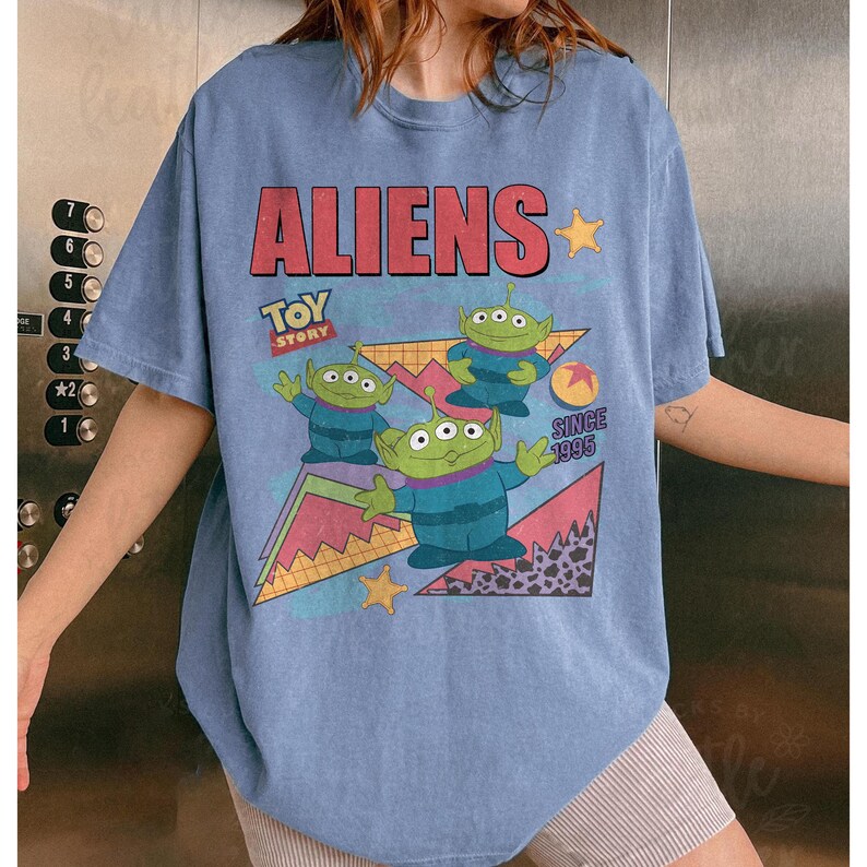May include: A light blue t-shirt featuring the word "ALIENS" in red block letters, with the "Toy Story" logo and three green alien characters. The shirt includes the text "SINCE 1995" and star graphics.