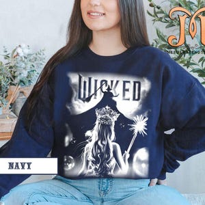May include: Navy blue sweatshirt featuring a graphic from the musical "Wicked." The design includes the word "Wicked" above a silhouette of a witch and a princess with a crown and wand. The word "NAVY" is printed at the bottom.