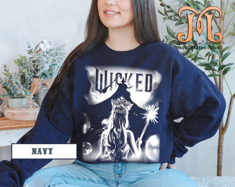 May include: Navy blue sweatshirt featuring a graphic from the musical "Wicked." The design includes the word "Wicked" above a silhouette of a witch and a princess with a crown and wand. The word "NAVY" is printed at the bottom.