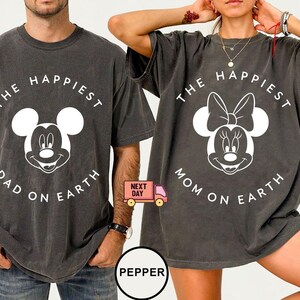 May include: Two charcoal gray t-shirts with white graphic designs. One shirt features Mickey Mouse with the text "THE HAPPIEST DAD ON EARTH." The other shirt features Minnie Mouse with the text "THE HAPPIEST MOM ON EARTH."