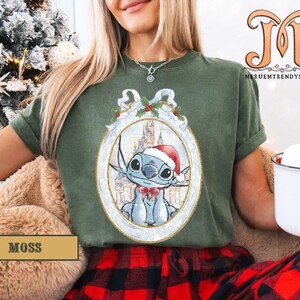 May include: Moss green t-shirt featuring a festive Stitch graphic in a Christmas ornament design. The graphic includes Stitch wearing a Santa hat, with a castle in the background. The shirt is part of a holiday collection.