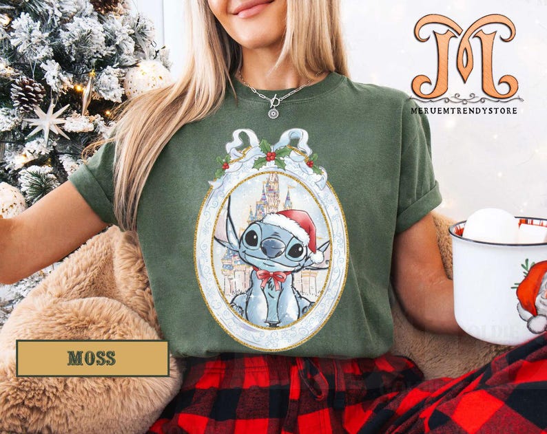 May include: Moss green t-shirt featuring a festive Stitch graphic in a Christmas ornament design. The graphic includes Stitch wearing a Santa hat, with a castle in the background. The shirt is part of a holiday collection.