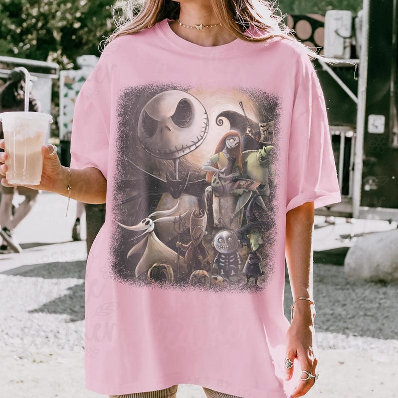 May include: A light pink t-shirt featuring a graphic from "The Nightmare Before Christmas." The design includes Jack Skellington, Sally, Zero, and other characters. The artwork is set against a dark, textured background.