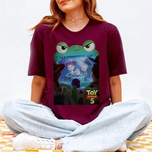 May include: Burgundy t-shirt with a graphic from Disney Pixar's Toy Story 5. The design features Woody, Buzz Lightyear, Jessie, and other characters. The shirt has a crew neck and short sleeves.