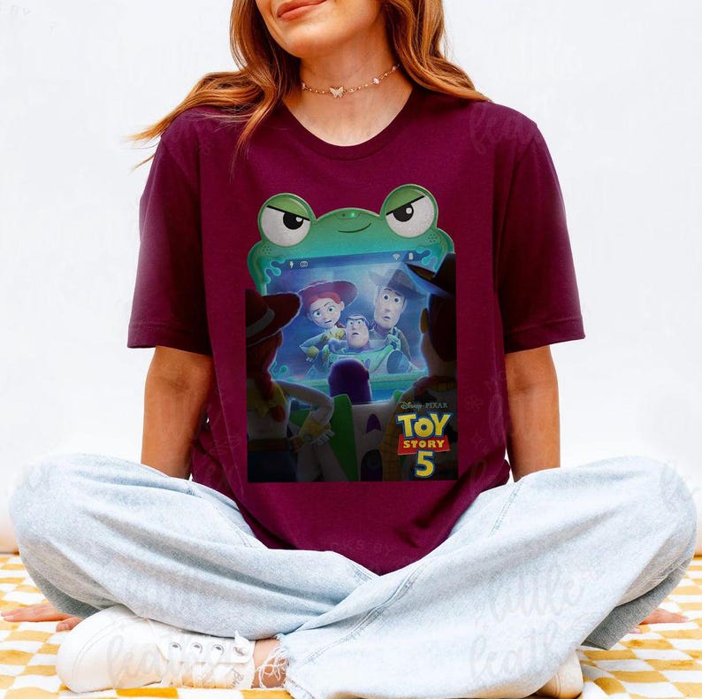 May include: Burgundy t-shirt with a graphic from Disney Pixar's Toy Story 5. The design features Woody, Buzz Lightyear, Jessie, and other characters. The shirt has a crew neck and short sleeves.