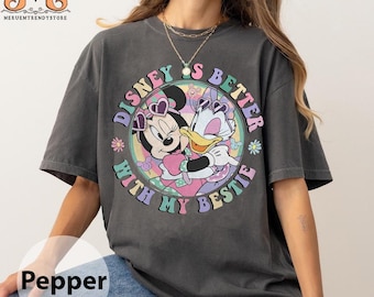 Op de afbeelding: Een grijs T-shirt met een kleurrijke afbeelding van Minnie Mouse en Daisy Duck die elkaar omhelzen. De afbeelding is omgeven door een cirkel met de tekst "Disney is better with my bestie".