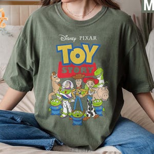 May include: Moss green t-shirt featuring the Disney Pixar Toy Story logo and characters. The design includes Woody, Buzz Lightyear, Jessie, and other characters from the animated film. The shirt is a relaxed fit.