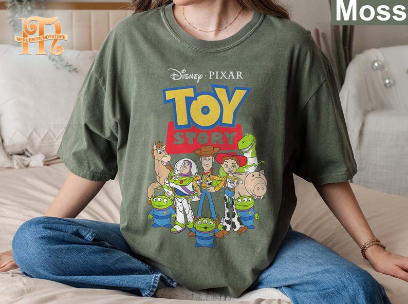 May include: Moss green t-shirt featuring the Disney Pixar Toy Story logo and characters. The design includes Woody, Buzz Lightyear, Jessie, and other characters from the animated film. The shirt is a relaxed fit.