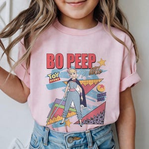 May include: Pink t-shirt featuring a graphic of Bo Peep from Toy Story. The design includes the text "BO PEEP" in red, "TOY STORY" in blue, and "little" in white. The shirt also has a retro geometric background.
