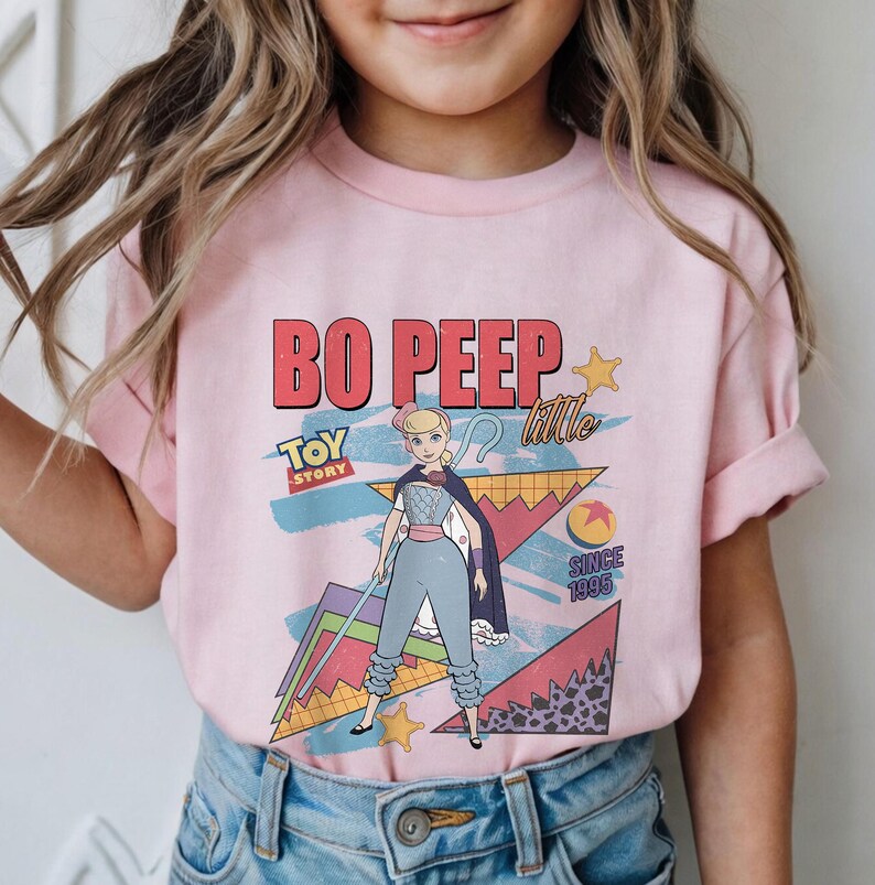 May include: Pink t-shirt featuring a graphic of Bo Peep from Toy Story. The design includes the text "BO PEEP" in red, "TOY STORY" in blue, and "little" in white. The shirt also has a retro geometric background.