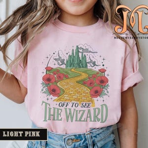 Puede incluir: Camiseta rosa claro con un gráfico colorido del Camino de baldosas amarillas que conduce a la Ciudad Esmeralda. El diseño incluye amapolas rojas y el texto "OFF TO SEE THE WIZARD".
