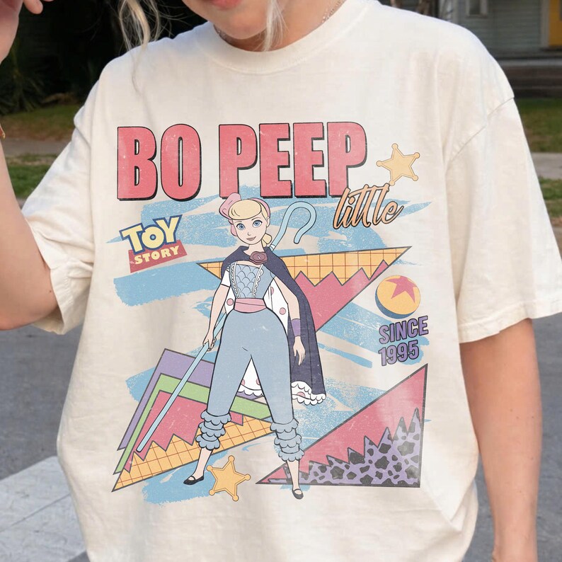 May include: Cream-colored t-shirt featuring a graphic of Bo Peep from Toy Story. The design includes the text "BO PEEP" in pink and red, "TOY STORY" logo, and "SINCE 1995". Bo Peep is depicted in her signature outfit with a staff.