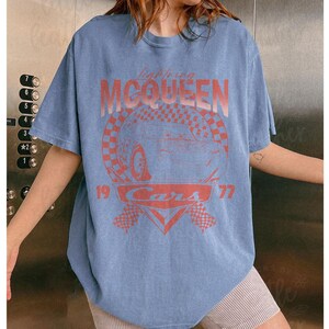 May include: A light blue t-shirt featuring a graphic of Lightning McQueen from the movie Cars. The design includes the text "Lightning McQueen," "Cars," and the numbers "19" and "77" in a red-orange color. The shirt has short sleeves.