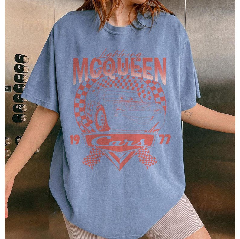 May include: A light blue t-shirt featuring a graphic of Lightning McQueen from the movie Cars. The design includes the text "Lightning McQueen," "Cars," and the numbers "19" and "77" in a red-orange color. The shirt has short sleeves.