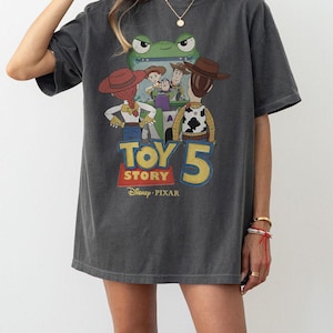 May include: Dark gray t-shirt featuring a graphic of characters from the animated movie Toy Story. The design includes the text "TOY STORY 5" in yellow and red, with the Disney Pixar logo.