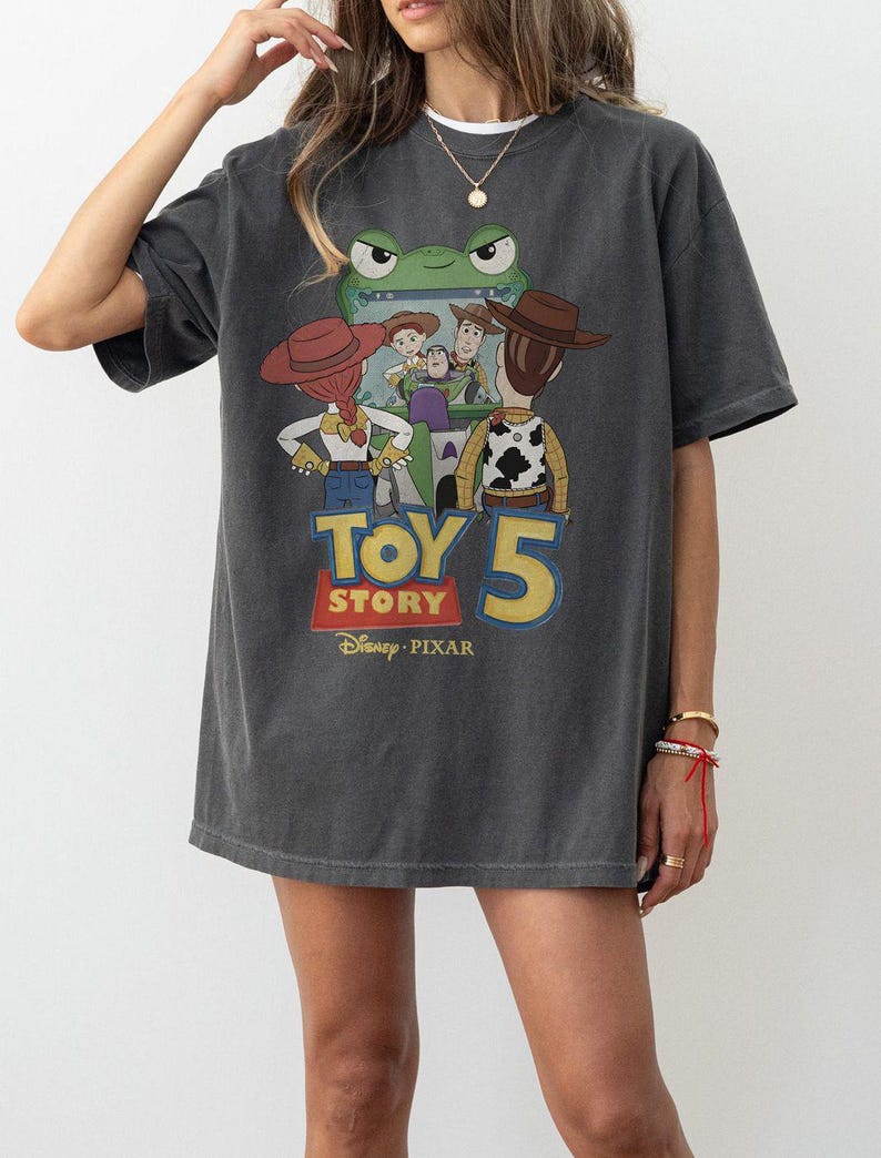 May include: Dark gray t-shirt featuring a graphic of characters from the animated movie Toy Story. The design includes the text "TOY STORY 5" in yellow and red, with the Disney Pixar logo.