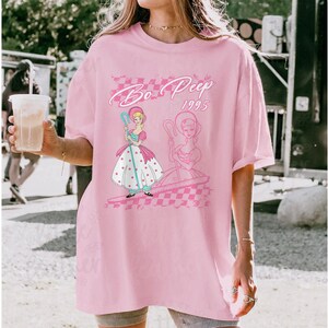 K&ouml;nnte beinhalten: Hellrosa T-Shirt mit einem Aufdruck von Bo Peep aus Toy Story. Das Design enth&auml;lt den Text "Bo Peep 1995" in einer Schreibschrift, zusammen mit einer Illustration von Bo Peep in einem rosa und wei&szlig;en Kleid. Das T-Shirt hat einen Rundhalsausschnitt und kurze &Auml;rmel.