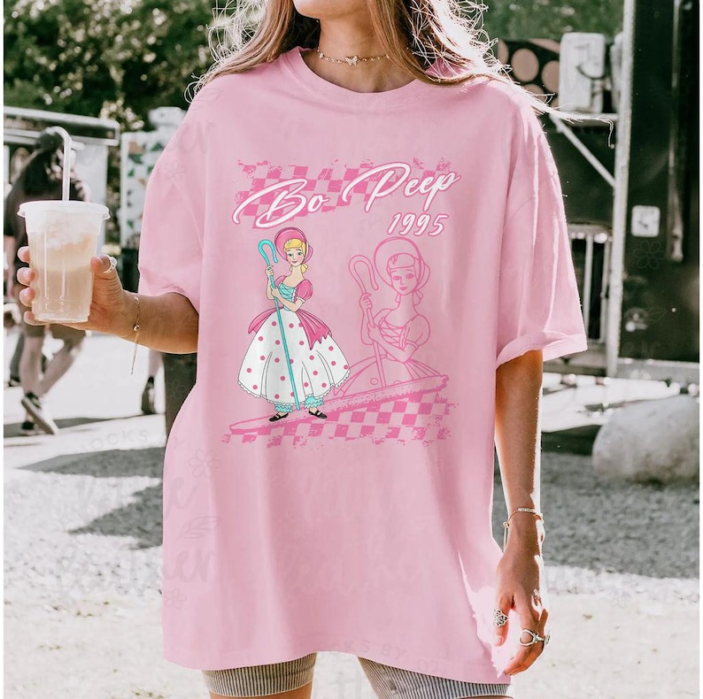 K&ouml;nnte beinhalten: Hellrosa T-Shirt mit einem Aufdruck von Bo Peep aus Toy Story. Das Design enth&auml;lt den Text "Bo Peep 1995" in einer Schreibschrift, zusammen mit einer Illustration von Bo Peep in einem rosa und wei&szlig;en Kleid. Das T-Shirt hat einen Rundhalsausschnitt und kurze &Auml;rmel.