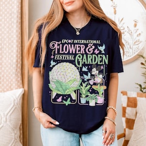 May include: Navy blue t-shirt featuring the text "EPCOT International Flower & Garden Festival" with a graphic of Spaceship Earth, Mickey Mouse, and various plants and butterflies. The design is in shades of pink, green, and blue.