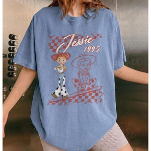 K&ouml;nnte beinhalten: Hellblaues T-Shirt mit einem Aufdruck der Figur Jessie aus dem Film Toy Story. Das Design enth&auml;lt den Text "Jessie 1995" und ein Schachbrettmuster. Das Shirt hat kurze &Auml;rmel.