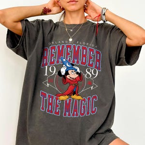 K&ouml;nnte beinhalten: Dunkelgraues T-Shirt mit Mickey Mouse als Zauberer. Der Text "REMEMBER THE MAGIC 1989 ORLANDO FLORIDA" ist rot und blau. Vintage-Design.