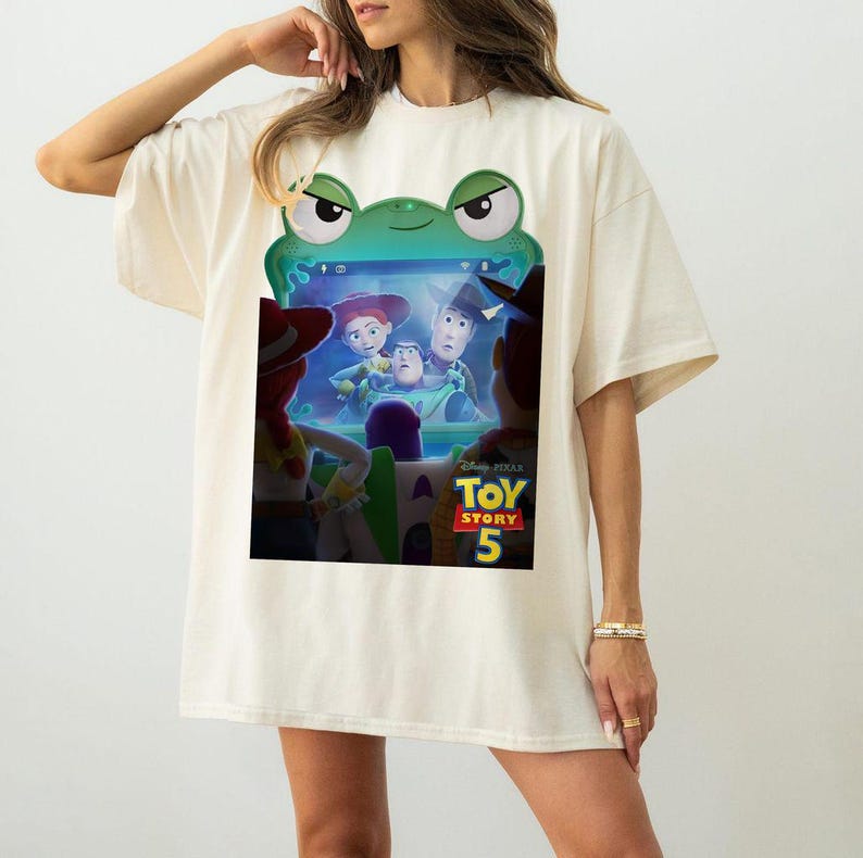 May include: Cream-coloured t-shirt featuring a green frog-shaped frame with a colourful graphic from the movie Toy Story 5. The graphic includes characters like Woody, Buzz Lightyear, and Jessie. The shirt is a casual, oversized fit.