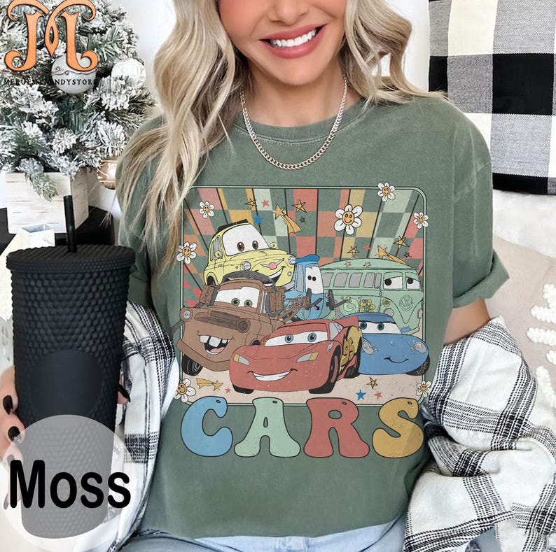 May include: A green t-shirt with a retro-style graphic of cartoon cars from the movie Cars. The graphic features a yellow car, a red car, a blue car, a brown car, and a green van. The text "CARS" is printed in colorful letters below the graphic.