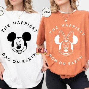 May include: Two t-shirts, one white and one orange, featuring Disney characters. The white shirt has Mickey Mouse with "THE HAPPIEST DAD ON EARTH" text. The orange shirt has Minnie Mouse with "THE HAPPIEST MOM ON EARTH" text.