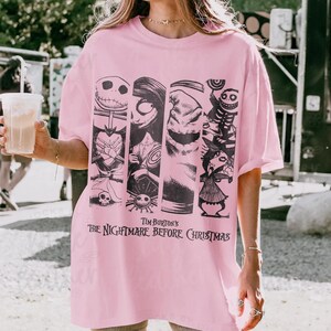 May include: A light pink t-shirt featuring a black graphic print of characters from Tim Burton's The Nightmare Before Christmas. The design includes vertical panels showing Jack Skellington, Sally, Oogie Boogie, and other figures from the film.