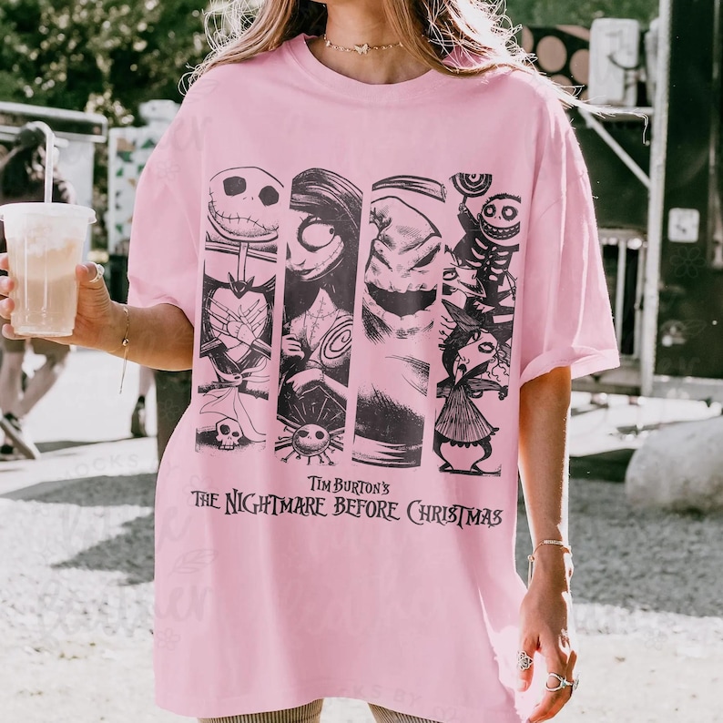 May include: A light pink t-shirt featuring a black graphic print of characters from Tim Burton's The Nightmare Before Christmas. The design includes vertical panels showing Jack Skellington, Sally, Oogie Boogie, and other figures from the film.