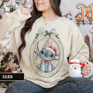 May include: Sand-colored sweatshirt featuring a framed illustration of Stitch wearing a Santa hat and a red bow tie. The illustration includes a castle in the background. The word "SAND" is printed on a black rectangle at the bottom.