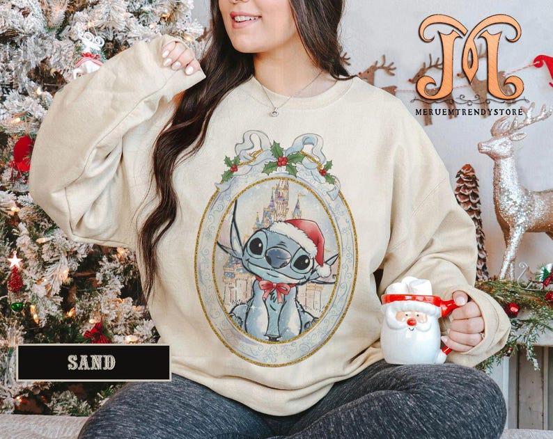 May include: Sand-colored sweatshirt featuring a framed illustration of Stitch wearing a Santa hat and a red bow tie. The illustration includes a castle in the background. The word "SAND" is printed on a black rectangle at the bottom.