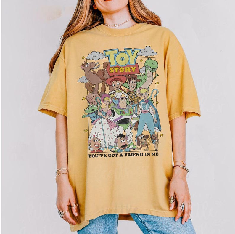 Puede incluir: Camiseta de color amarillo mostaza con un gr&aacute;fico colorido de los personajes de "Toy Story". El dise&ntilde;o incluye el t&iacute;tulo de la pel&iacute;cula y la frase "You've Got a Friend in Me."