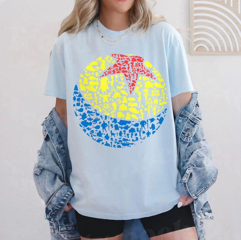 May include: Light blue t-shirt featuring a graphic of a yellow, red, and blue ball. The ball design is filled with character silhouettes. The shirt is made of a soft material.