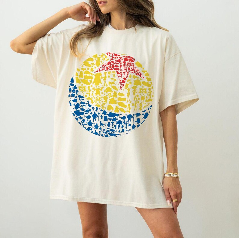May include: Cream-colored t-shirt featuring a large, colorful graphic of a ball. The graphic is a circle divided into sections of red, yellow, and blue, filled with various character silhouettes. The shirt has short sleeves and a relaxed fit.