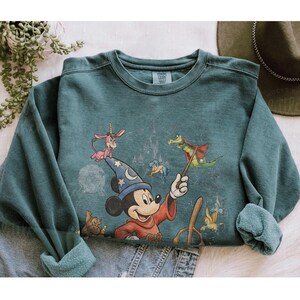 K&ouml;nnte beinhalten: T&uuml;rkisfarbenes Sweatshirt mit einer Grafik von Mickey Mouse als Zauberer, umgeben von Zeichentrickfiguren. Das Design umfasst ein Schloss und andere skurrile Elemente. Das Sweatshirt hat einen Rundhalsausschnitt und lange &Auml;rmel.