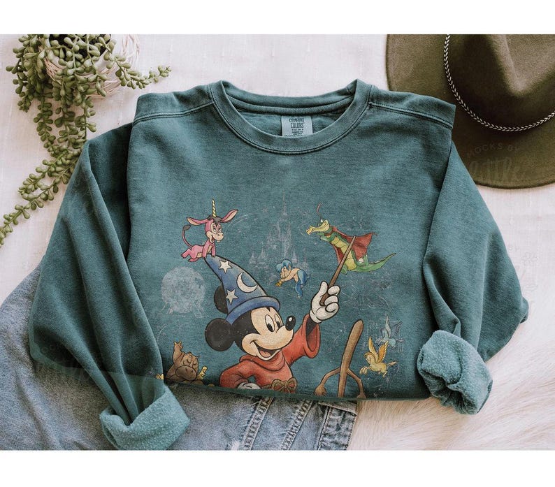 K&ouml;nnte beinhalten: T&uuml;rkisfarbenes Sweatshirt mit einer Grafik von Mickey Mouse als Zauberer, umgeben von Zeichentrickfiguren. Das Design umfasst ein Schloss und andere skurrile Elemente. Das Sweatshirt hat einen Rundhalsausschnitt und lange &Auml;rmel.