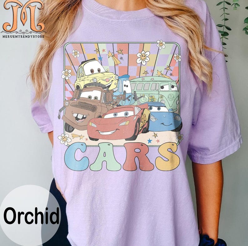 May include: A purple t-shirt with a retro-style graphic of cartoon cars from the Disney Pixar movie Cars. The graphic features Lightning McQueen, Mater, and other characters. The text "CARS" is printed in rainbow colors below the graphic.