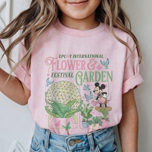 May include: Pink t-shirt featuring the text "Epcot International Flower & Garden Festival". The design includes a large floral sphere, Mickey Mouse watering plants, and butterflies. A fun and colorful tee for kids.