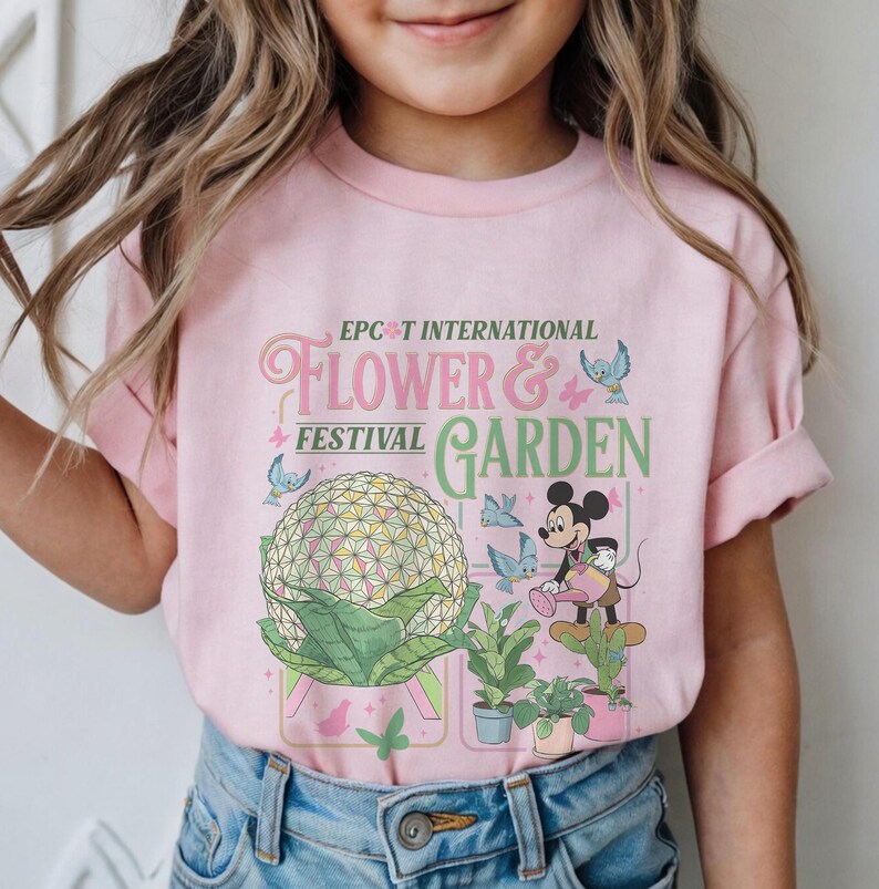 May include: Pink t-shirt featuring the text "Epcot International Flower & Garden Festival". The design includes a large floral sphere, Mickey Mouse watering plants, and butterflies. A fun and colorful tee for kids.