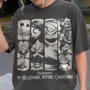 May include: A charcoal gray t-shirt featuring a graphic print of characters from Tim Burton's The Nightmare Before Christmas. The design consists of four vertical panels displaying Jack Skellington, Sally, Oogie Boogie, and other figures.