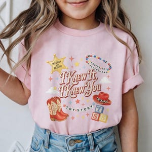 May include: A light pink t-shirt featuring a graphic print with the text "I KNEW IT, I KNEW YOU" and "TOY STORY". The design includes a yellow star, a beaded necklace, a red cowboy hat, a pair of red cowboy boots, and three colourful alphabet blocks.