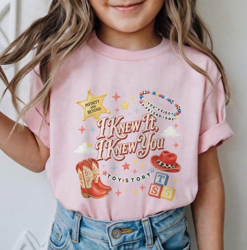 May include: A light pink t-shirt featuring a graphic print with the text "I KNEW IT, I KNEW YOU" and "TOY STORY". The design includes a yellow star, a beaded necklace, a red cowboy hat, a pair of red cowboy boots, and three colourful alphabet blocks.