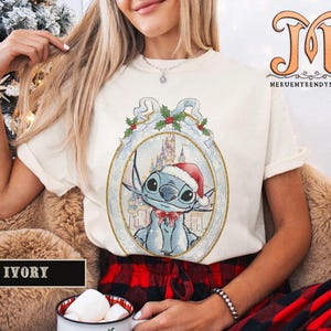 May include: Ivory-colored t-shirt featuring a cartoon Stitch character wearing a Santa hat and a red bow tie, set against a winter castle backdrop within a decorative frame. The shirt has short sleeves and a crew neck.