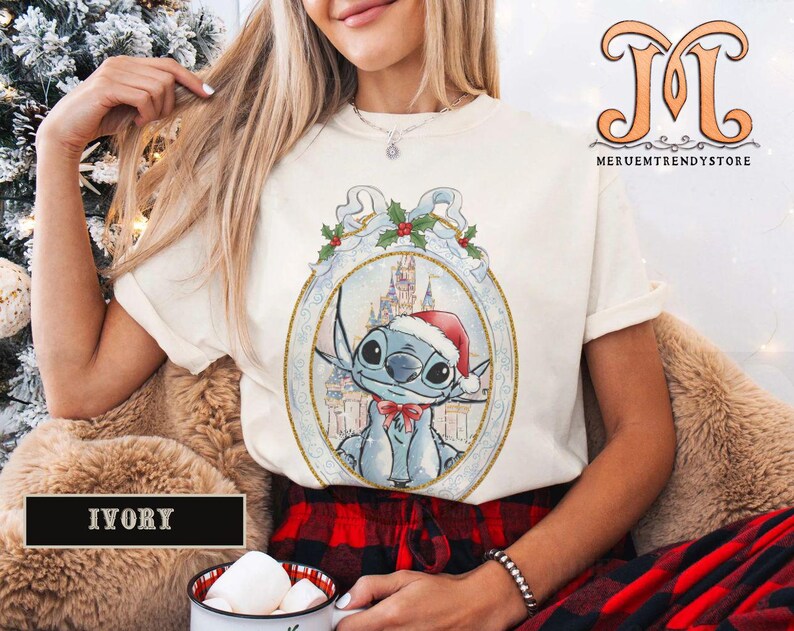May include: Ivory-colored t-shirt featuring a cartoon Stitch character wearing a Santa hat and a red bow tie, set against a winter castle backdrop within a decorative frame. The shirt has short sleeves and a crew neck.