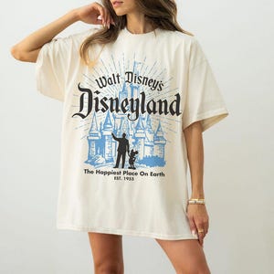Puede incluir: Camiseta color crema con el texto "Walt Disney's Disneyland" en escritura negra sobre un gr&aacute;fico azul de un castillo y siluetas de una persona y Mickey Mouse. El texto "The Happiest Place On Earth" y "EST. 1955" est&aacute;n debajo.